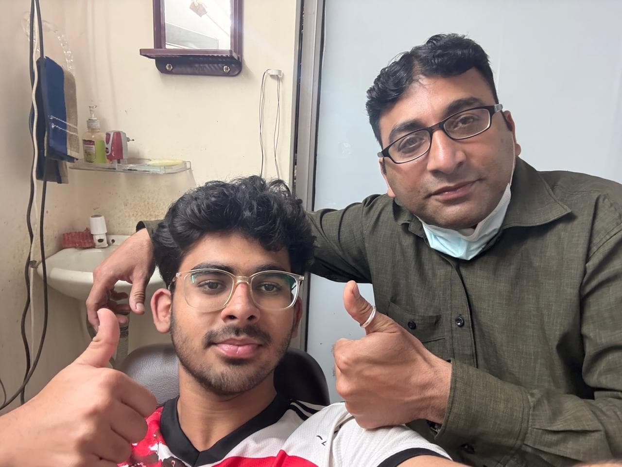 Two men wearing glasses give a thumbs up while posing together in a small room.